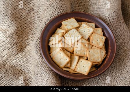 Salty Crackers in a clay bowl on burlap. Top view of a full bowl of square treats. Close-up. selective focus. Stock Photo