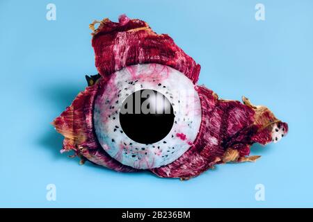 Dragon Fruit or Pitaya half peeled from the skin with black circle depicting eye pupil on blue background.