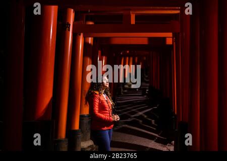 Kyoto, Japan with young caucasian woman standing by red Fushimi Inari shrine torii gates in park at night with illumination lanterns by path Stock Photo