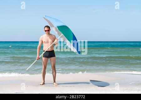 Fit muscular man on ocean beach shore of Santa Rosa, Panhandle Florida with sunglasses holding umbrella with waves crashing in background Stock Photo