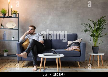 Worried man sitting on couch at home. Frustrated confused man feels unhappy, problems in personal life. Stock Photo
