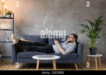 Young man with Glasses is Lying on the Couch and Texting on the Phone. Stock Photo