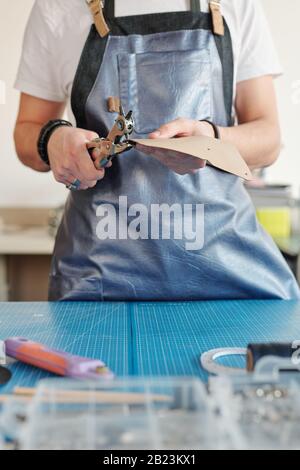 Young creative man in workwear standing by table while holding piece of light leather and making holes Stock Photo