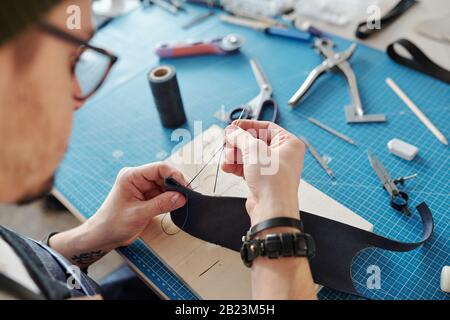 Contemporary craftsman sewing two parts of black leather together while holding them over wooden board on table Stock Photo