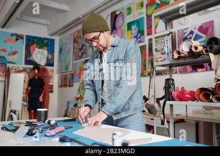 Young handicraft master in beanie hat, denim jacket and jeans outlining paper pattern by table in his workshop Stock Photo
