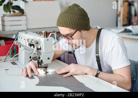 Serious young male leatherworker bending over electric sewing machine while holding piece of leather under needle during work Stock Photo