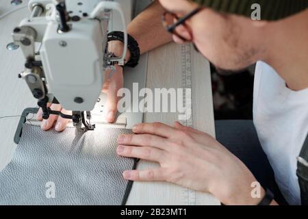 Hands of young leatherworker sewing zipper and piece of leather together while bending over electric machine in workshop Stock Photo