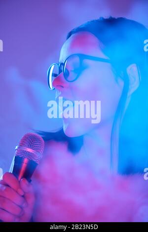 Pretty young emotional female singer with microphone by mouth performing songs in smoke against neon background Stock Photo