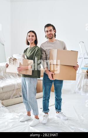 Cheerful young couple in casualwear holding carton boxes with packed stuff in living-room of their new house Stock Photo