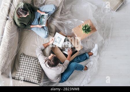Top view of young female with touchpad sitting on couch while her husband unpacking one of boxes near by Stock Photo