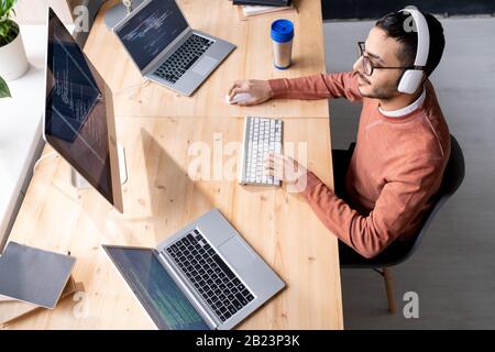 Busy young software developer in headphones sitting by table in front of two laptops and computer monitor during work Stock Photo