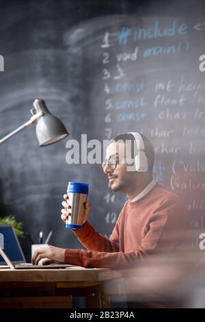 Serious busy manager or programmer in headphones having drink while working by laptop and listening to music Stock Photo