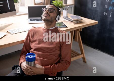 Young restful programmer in headphones holding mug with drink while sitting on chair against his workplace at break Stock Photo