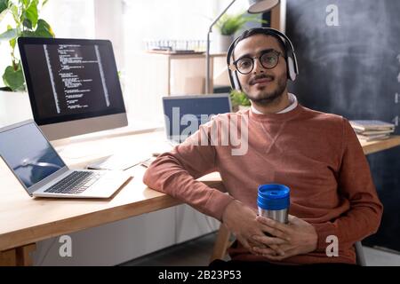 Young handsome programmer in headphones listening to music and having drink while sitting on chair by desk at break Stock Photo