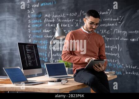 Clever young manager using touchpad while sitting on table with two laptops and computer monitor with coded data Stock Photo