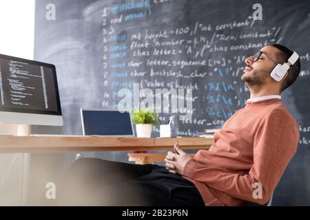 Young relaxed office manager or programmer enjoying music in headphones while sitting against blackboard Stock Photo