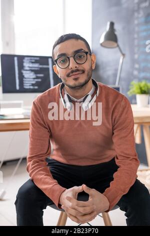 Young casual programmer with headphones looking at you while sitting in front of camera with workplace on background Stock Photo