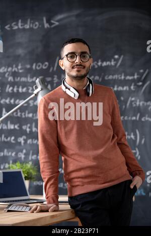 Young confident it-manager in casualwear and eyeglasses standing by desk with laptop on background of blackboard with formula Stock Photo