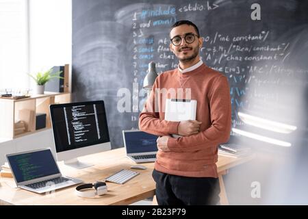 Young casual software developer with digital tablet standing by table in front of camera with blackboard on background Stock Photo