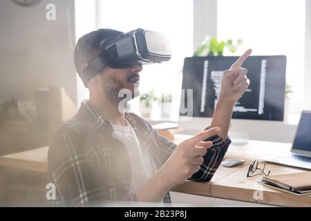 Contemporary software developer in vr headset pointing at virtual display while making presentation of new program Stock Photo