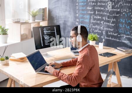 Busy young programmer with headphones analyzing data on laptop display and computer screen while sitting by workplace Stock Photo