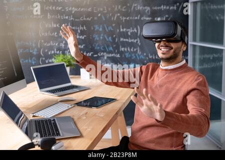 Happy young programmer wearing vr goggle sitting by desk with laptops and looking at virtual display Stock Photo