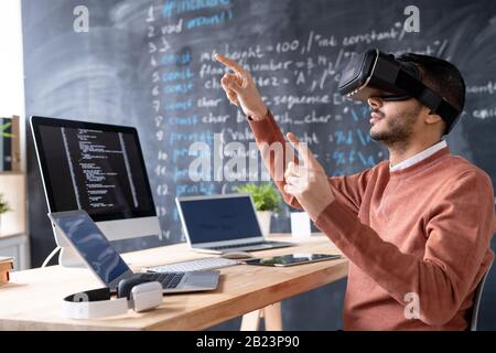 Young programmer in vr headset testing or presenting new software while pointing at virtual display by workplace Stock Photo