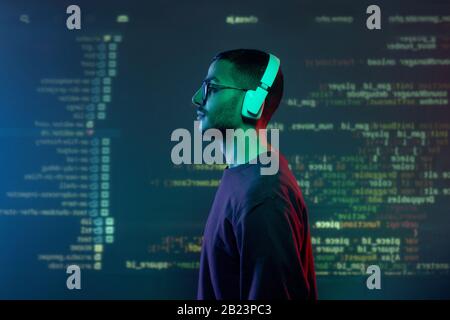 Side view of young programmer in headphones and casualwear standing against screen with decoded information Stock Photo