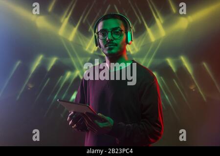 Young serious software developer in headphones using touchpad while standing in front of camera in neon lights Stock Photo