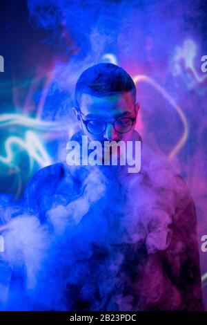 Head of young man in eyeglasses standing in isolation in front of camera among smoke and blue neon light Stock Photo