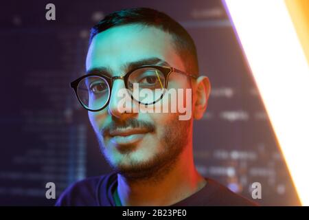 Face of young clever confident programmer or software developer in eyeglasses standing against large screen with data Stock Photo