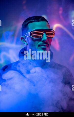 Head of contemporary young mixed-race man in red spiral eyewear among smoke and blue neon light Stock Photo