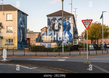 Two murals face each other in the Glenfada Park pedestrian area in the Bogside district of Londonderry, County Derry, Northern Ireland: on the left, a blue-toned figure in a sweeping motion; on the right, several fleeing figures shielding their faces above a memorial plinth reading "Stand by the Republic." Glenfada Park North was one of the sites where British soldiers shot civilians on "Bloody Sunday" in 1972. Stock Photo