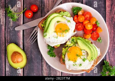 Avocado toasts with eggs and tomatoes on whole grain bread. Above view on a dark wood background. Stock Photo
