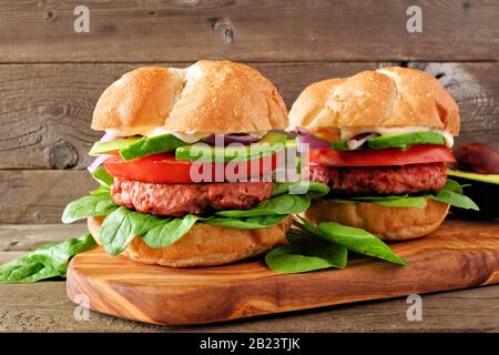 Plant based meatless burgers with avocado, tomato and spinach on a serving board against a rustic wood background Stock Photo
