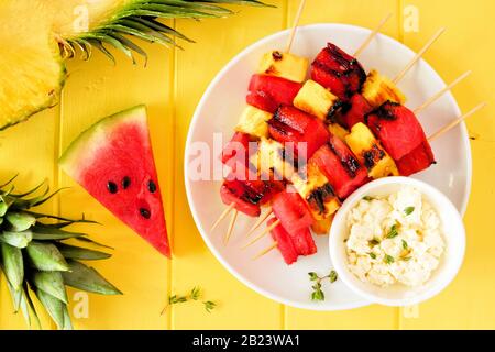 Grilled watermelon and pineapple fruit skewers with feta. Above view on a bright yellow wood background. Summer food concept. Stock Photo