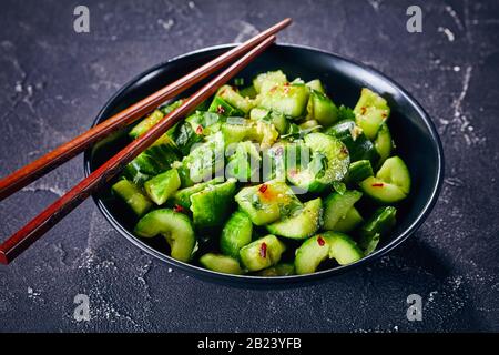 Smashed Cucumber salad sprinkled with fresh coriander leaves in a black bowl with chopsticks on a concrete table, close-up, Stock Photo