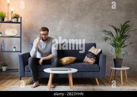 Young man in glasses angry talking on the phone sitting on the couch. Young businessman. Stock Photo