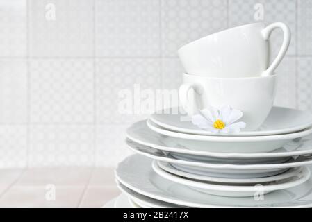 Stack of white porcelain plates and tea cups are on the kitchen table against the background of a apron made of ceramic tile, with copy-space Stock Photo