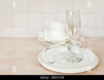 Stack of white porcelain plates, tea cups and wine glasses on the kitchen table against the background of a apron made of ceramic tile Stock Photo