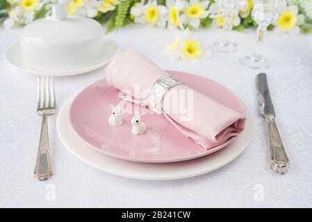 Classic serving for a Easter dinner with white and pink porcelain plates, pink napkin, silverware, two Easter rabbits and spring flowers are on a whit Stock Photo