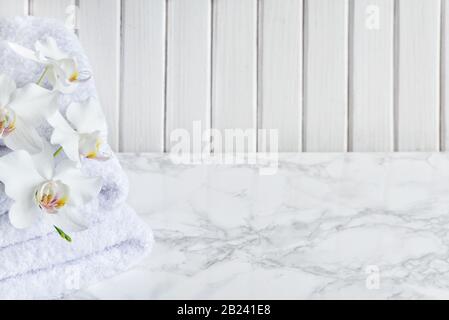 Stack of white terry towels and white orchid flowers are on a marble surface against a white background of wooden planks, with copy-space Stock Photo