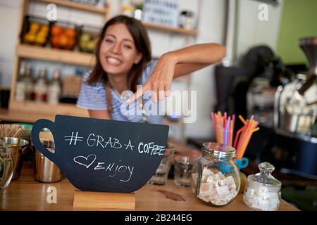 Smiling woman owner shows grab sign for coffee. Stock Photo