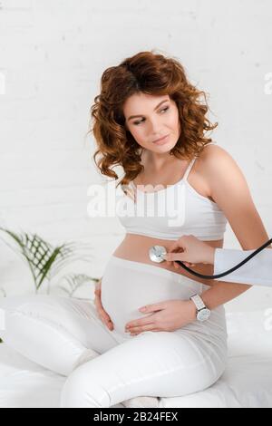Beautiful pregnant woman sitting on massage table during doctor examining belly with stethoscope in clinic Stock Photo