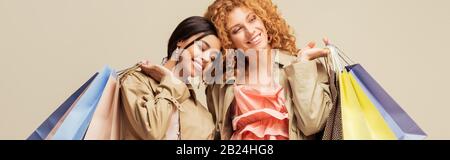 panoramic shot of happy multicultural girls in trench coats holding shopping bags isolated on beige Stock Photo