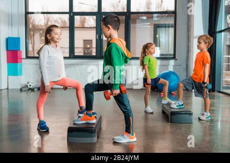 Selective focus of multiethnic children holding dumbbells and working out together on step platforms in gym Stock Photo