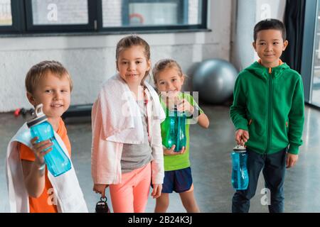 Happy multicultural children with towels holding sports bottles in gym Stock Photo