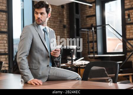 confident, handsome businessman sitting on office desk, holding smartphone and looking away Stock Photo