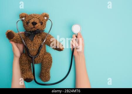 Cropped view of woman holding teddy bear with stethoscope on blue background, international childhood cancer day concept Stock Photo