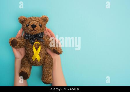 Cropped view of woman holding teddy bear with yellow ribbon on blue background, international childhood cancer day concept Stock Photo